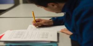 Student wearing blue sweatshirt taking notes with a pencil in a classroom