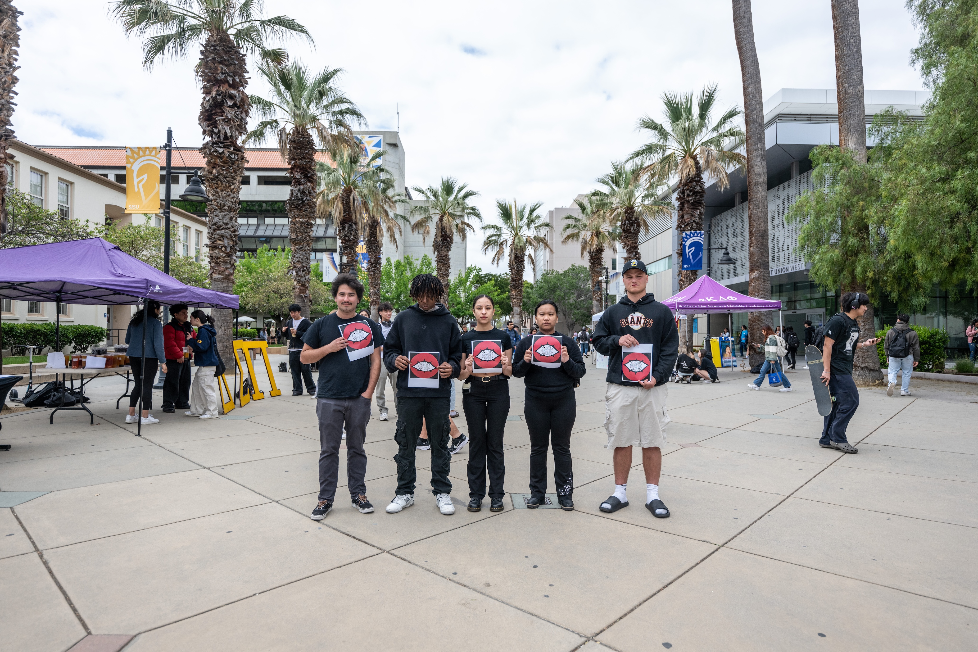 5 people standing on paseo holding 1984 graphics with palm trees behind them on a cloudy day