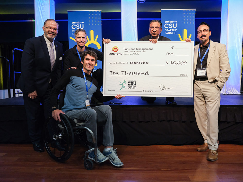 A male, college-age startup founder in a wheelchair poses with two sponsors, two hosts, and a giant check for $25,000