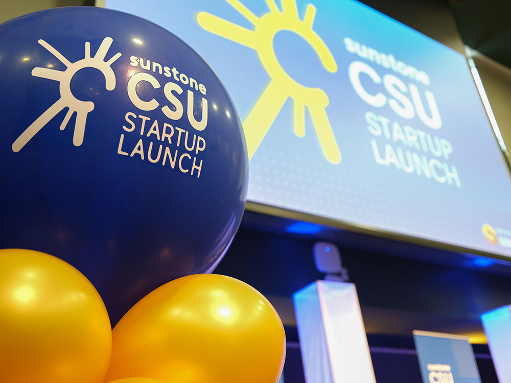 A blue balloon with a starburst logo and the words "Sunstone CSU Startup Competition" in the foreground, with a screen on which is projected the same logo on a blue background behind it.