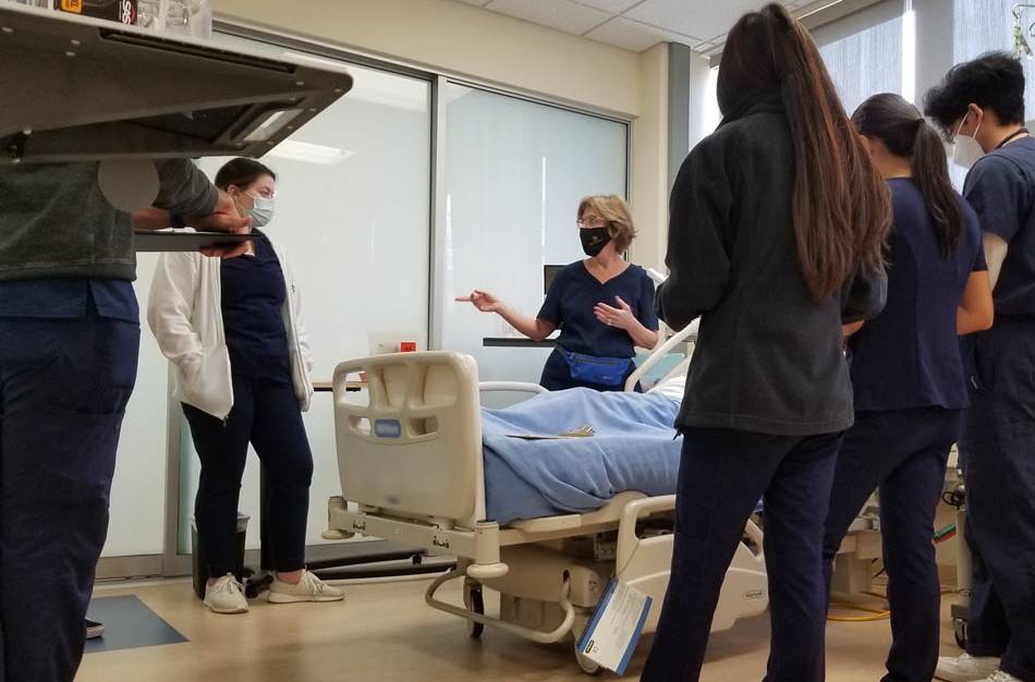 TVFSON nursing instructor leading a class of students, all wearing blue scrubs, in the nursing simulation lab. Photo Credit: Maya Carlyle, 2021 November.